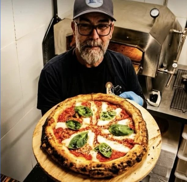 Francis Douglas segurando uma pizza margherita na pá, em frente ao forno